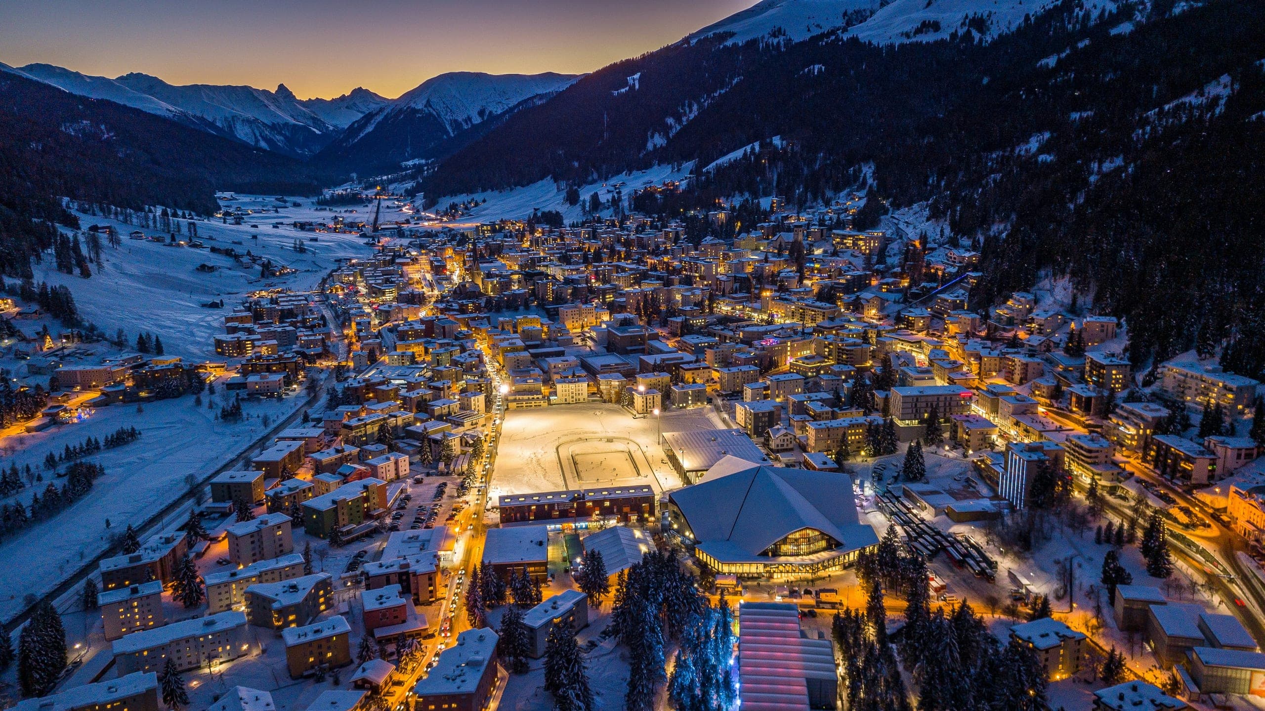 An aerial view of Davos at dusk, lit windows across the valley beneath snow-covered peaks