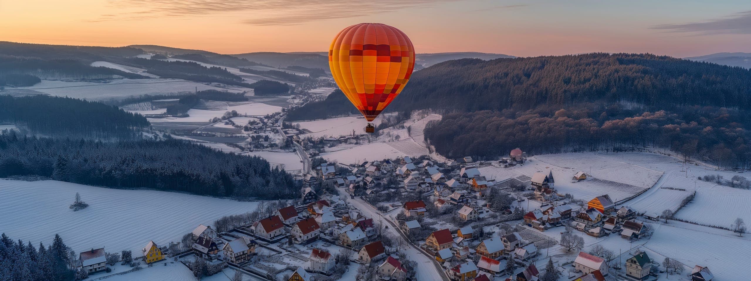A hot-air balloon drifting at dawn over a snow-covered alpine village and forested hills