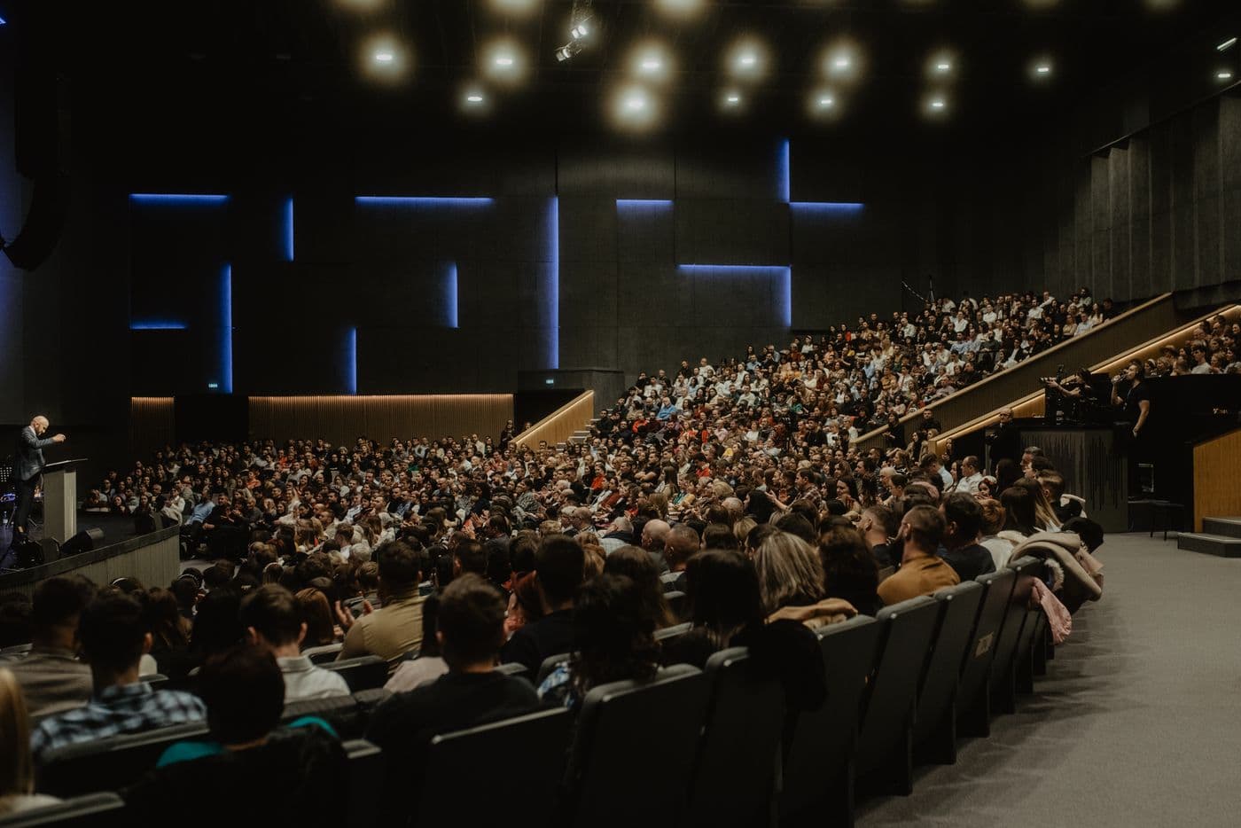 A speaker at a lectern addressing a full tiered auditorium lit in blue